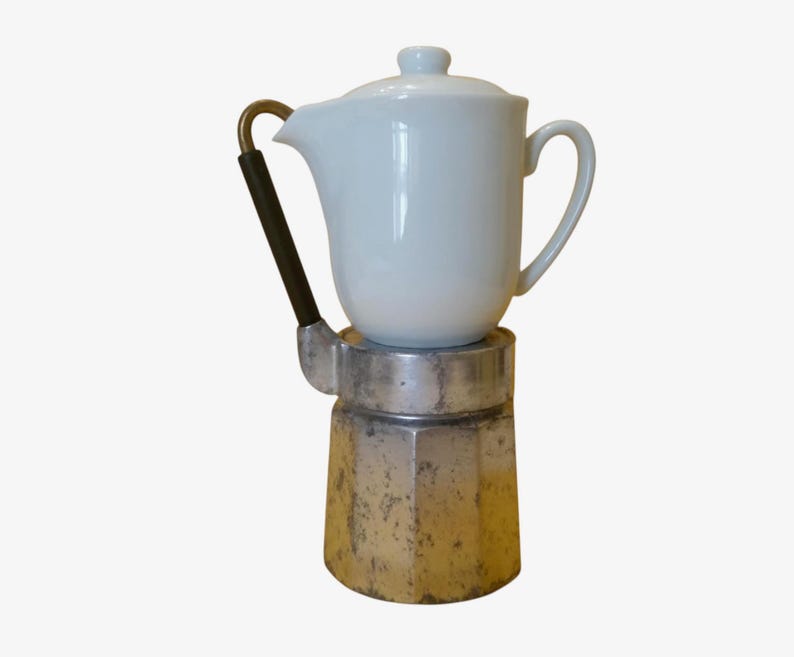 May include: A vintage stovetop espresso maker with a white ceramic top and a silver-colored base. The handle is black with a brass accent. The coffee maker is designed to brew espresso on a stovetop.