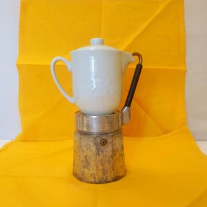 May include: A white ceramic coffee pot with a lid sits atop a vintage, weathered metal base. The pot has a curved handle and a black handle on the side. The background is a bright yellow cloth.