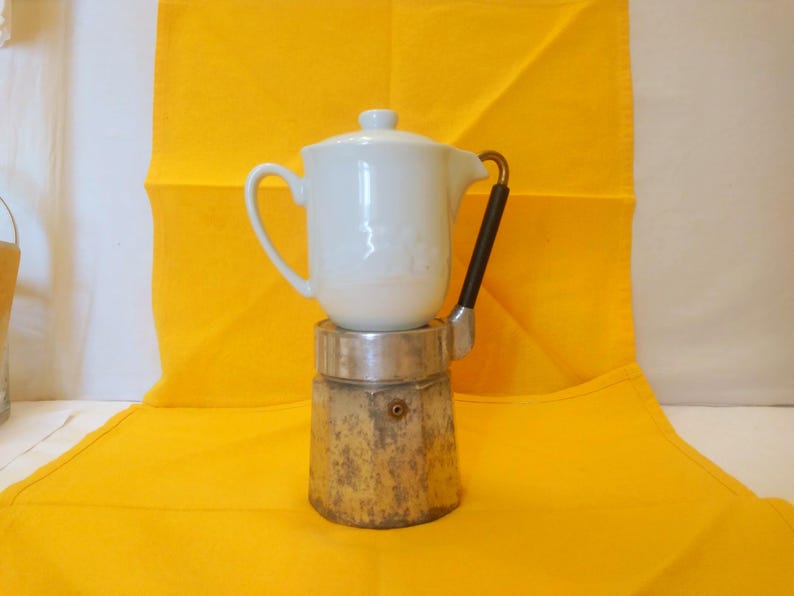 May include: A white ceramic coffee pot with a lid sits atop a vintage, weathered metal base. The pot has a curved handle and a black handle on the side. The background is a bright yellow cloth.