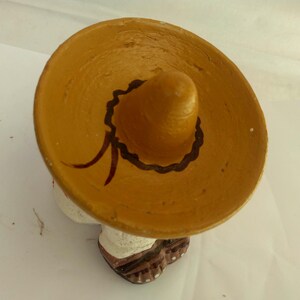 May include: A ceramic figurine of a person wearing a large, tan sombrero with a dark brown trim and red accents. The figurine has a white shirt and brown shoes. The figure is a traditional Mexican folk art piece.
