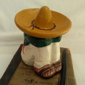 May include: A small, painted figurine of a person wearing a sombrero. The figure has a white body, green collar, and brown shoes. The sombrero is tan with a brown decorative border. The figurine is resting on a book with text.