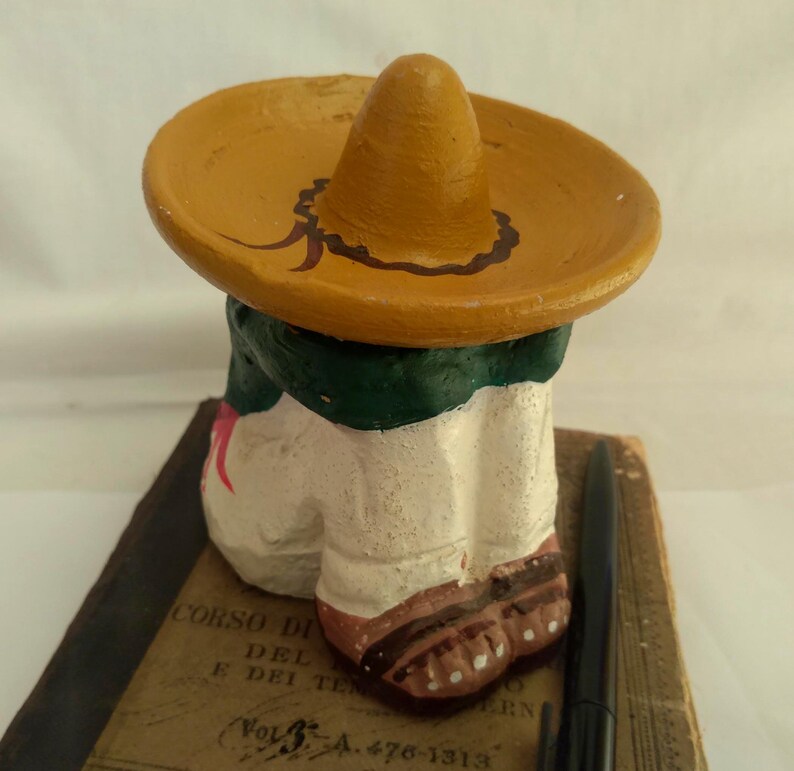 May include: A small, painted figurine of a person wearing a sombrero. The figure has a white body, green collar, and brown shoes. The sombrero is tan with a brown decorative border. The figurine is resting on a book with text.