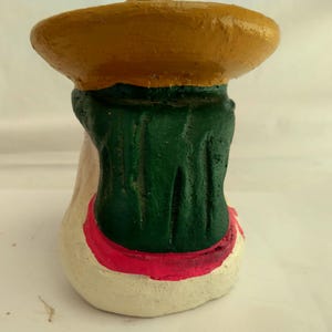 May include: A handcrafted, painted ceramic figurine. The figurine features a wide, yellow brimmed hat, a green torso, and a cream-colored base with a red trim. The figurine is a unique piece of art.
