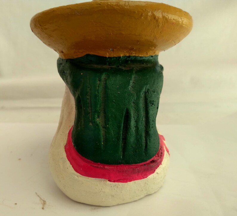 May include: A handcrafted, painted ceramic figurine. The figurine features a wide, yellow brimmed hat, a green torso, and a cream-colored base with a red trim. The figurine is a unique piece of art.