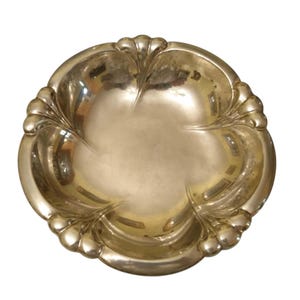 May include: A silver-toned decorative bowl with a scalloped edge and a four-lobed interior design. The bowl has a reflective surface and a detailed, ornate rim. The overall shape is round, with a shallow depth, suitable for display or serving.