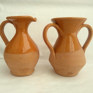 May include: Two antique terracotta pitchers with handles. The pitchers have a two-tone design, with a lighter, unglazed base and a glossy, orange-brown glazed upper portion. One pitcher has a spout, the other has a wide opening.