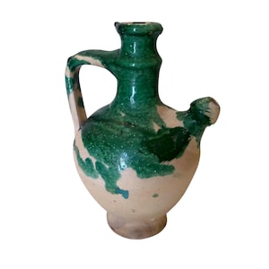 Puede incluir: Una jarra de cerámica con cuerpo redondeado y cuello estrecho, que presenta un asa en un lado y un pequeño pico vertedor en el otro. El recipiente tiene un color base beige claro con parches irregulares de esmalte verde oscuro en la parte superior.