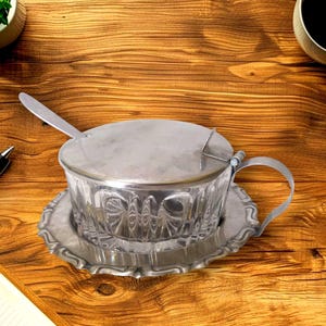 May include: A clear glass sugar bowl with a vertical ribbed pattern, featuring a silver-toned metal lid, a matching silver-toned metal base with a scalloped edge, a curved side handle, and a small silver-toned metal spoon resting inside.