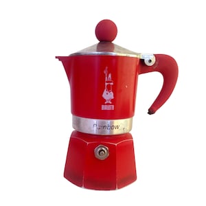 May include: A red Bialetti Rainbow stovetop espresso maker. The coffee pot has a silver band and a red handle and knob. The Bialetti logo is visible on the front. The word "Rainbow" is printed on the bottom.