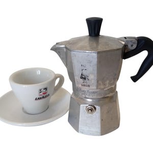 Pode incluir: Uma cafeteira de alumínio prateado com pega e botão da tampa pretos, apresentando um logótipo estampado e o texto Moka Express. Ao lado, encontra-se uma chávena de café em cerâmica branca com pires e o logótipo Lavazza impresso.