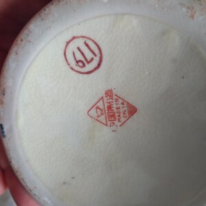 Pu&ograve; includere: Primo piano del fondo di un oggetto in ceramica, probabilmente un vaso o una ciotola. La base &egrave; bianco sporco con un timbro rosso circolare che indica "611". &Egrave; visibile anche un timbro rosso a forma di diamante con caratteri cinesi e "MADE IN CHINA".