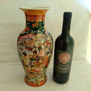Pu&ograve; includere: Un vaso in ceramica ornato, con bordo svasato e una scena dipinta di figure e paesaggi. Accanto c'&egrave; una bottiglia di vino rosso. Il vaso ha un bordo dorato e uno sfondo verde scuro con dettagli floreali. Il vaso &egrave; alto circa 30 cm.