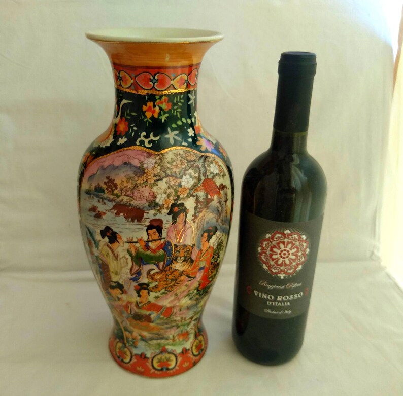 Pu&ograve; includere: Un vaso in ceramica ornato, con bordo svasato e una scena dipinta di figure e paesaggi. Accanto c'&egrave; una bottiglia di vino rosso. Il vaso ha un bordo dorato e uno sfondo verde scuro con dettagli floreali. Il vaso &egrave; alto circa 30 cm.