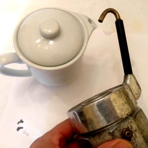 May include: A white ceramic teapot with a lid and handle is next to a vintage silver and black espresso maker. The espresso maker has a brass spout and a black handle. The items are on a white surface.