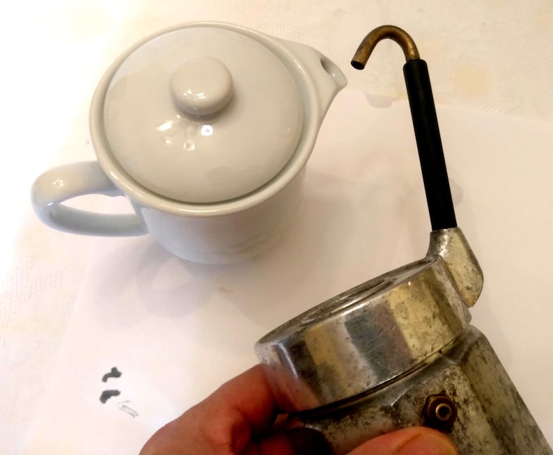 May include: A white ceramic teapot with a lid and handle is next to a vintage silver and black espresso maker. The espresso maker has a brass spout and a black handle. The items are on a white surface.