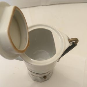 May include: White ceramic coffee pot with a lid, featuring a gold-colored handle and a decorative base. The lid is open, revealing the interior. The pot has a classic design, suitable for brewing coffee. The pot is likely used for serving coffee.