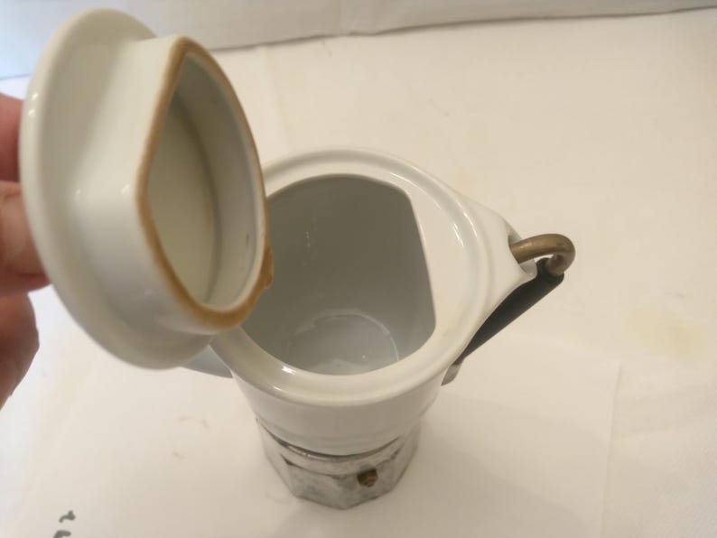 May include: White ceramic coffee pot with a lid, featuring a gold-colored handle and a decorative base. The lid is open, revealing the interior. The pot has a classic design, suitable for brewing coffee. The pot is likely used for serving coffee.