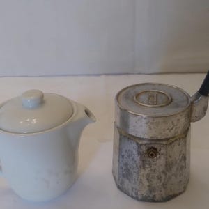 May include: A white ceramic teapot with a lid and a spout, next to a vintage silver stovetop espresso maker with a black handle and a curved spout. The espresso maker has a round top.