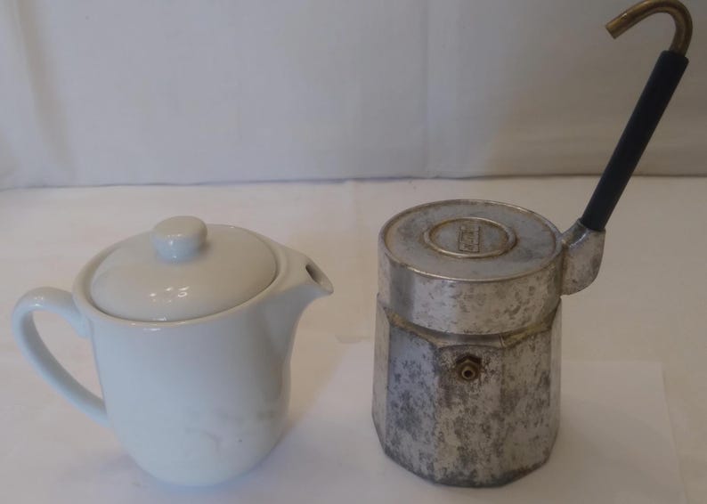 May include: A white ceramic teapot with a lid and a spout, next to a vintage silver stovetop espresso maker with a black handle and a curved spout. The espresso maker has a round top.
