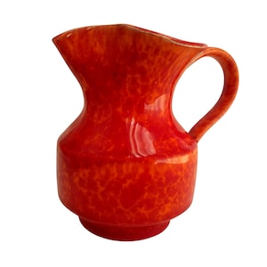 Puede incluir: Una jarra de cerámica roja vibrante con un patrón moteado. La jarra tiene un asa curva y un pico para verter fácilmente. La base es de un tono rojo ligeramente más oscuro. Un artículo de menaje decorativo.