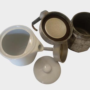 May include: An overhead shot of a disassembled stovetop espresso maker and a white ceramic creamer. The espresso maker is made of metal and has a black handle. The creamer has a handle and a spout.