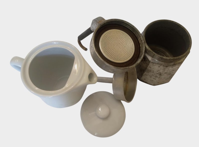 May include: An overhead shot of a disassembled stovetop espresso maker and a white ceramic creamer. The espresso maker is made of metal and has a black handle. The creamer has a handle and a spout.