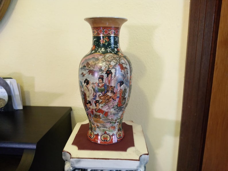 Pu&ograve; includere: Un vaso decorativo in ceramica con una scena dipinta dettagliata di figure e una tigre. Il vaso ha un bordo svasato e un corpo bulboso. I colori includono rosso, verde e oro. &Egrave; esposto su un supporto decorativo.