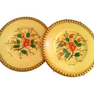 May include: Two decorative plates with a light yellow background and a gold beaded rim. Each plate features a hand-painted floral design with a red rose, green leaves, and orange buds. The plates have the word "gremmi" written on them.