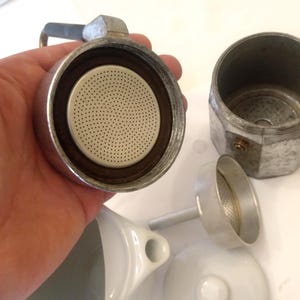 May include: Disassembled silver stovetop espresso maker with a perforated filter, a separate chamber, and a small funnel. The components are arranged on a white surface, showcasing the parts of the coffee brewing device.