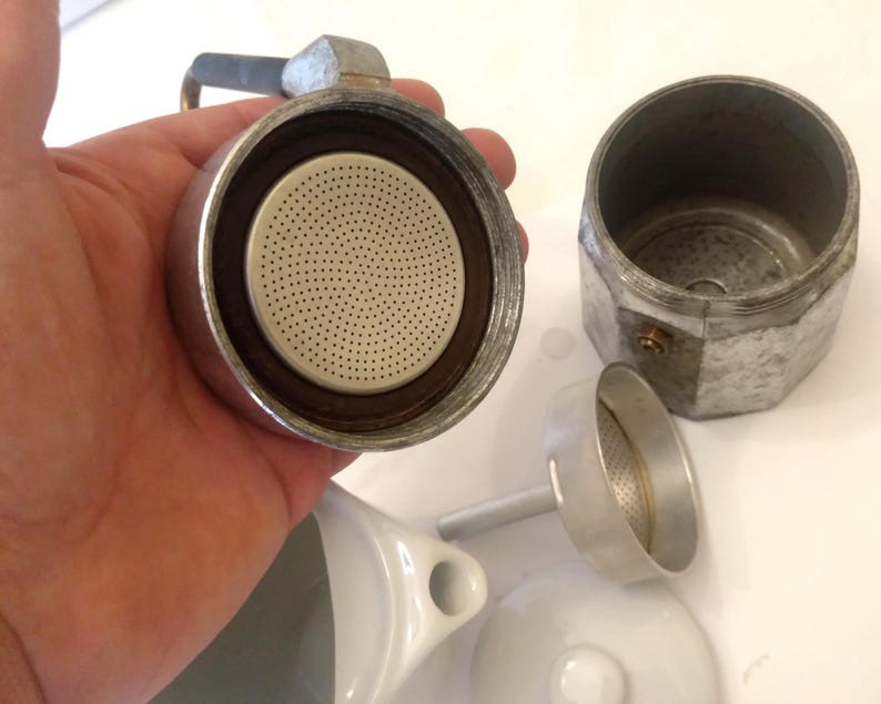 May include: Disassembled silver stovetop espresso maker with a perforated filter, a separate chamber, and a small funnel. The components are arranged on a white surface, showcasing the parts of the coffee brewing device.