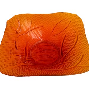 May include: Orange glass serving dish with a textured surface and leaf-like design. The dish has a square shape with rounded edges and a circular indentation in the center.
