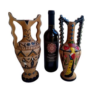 Op de afbeelding: Twee decoratieve amfora vazen met handvatten, naast een fles rode wijn. De vazen zijn bruin met zwart-witte geschilderde ontwerpen. De wijnfles heeft een zwart etiket met een bloemmotief.