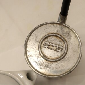 May include: Close-up of a vintage, circular metal object with a black tube attached. The object has a worn, brushed metal finish with a recessed center featuring a logo. A white ceramic object is visible in the lower left corner.