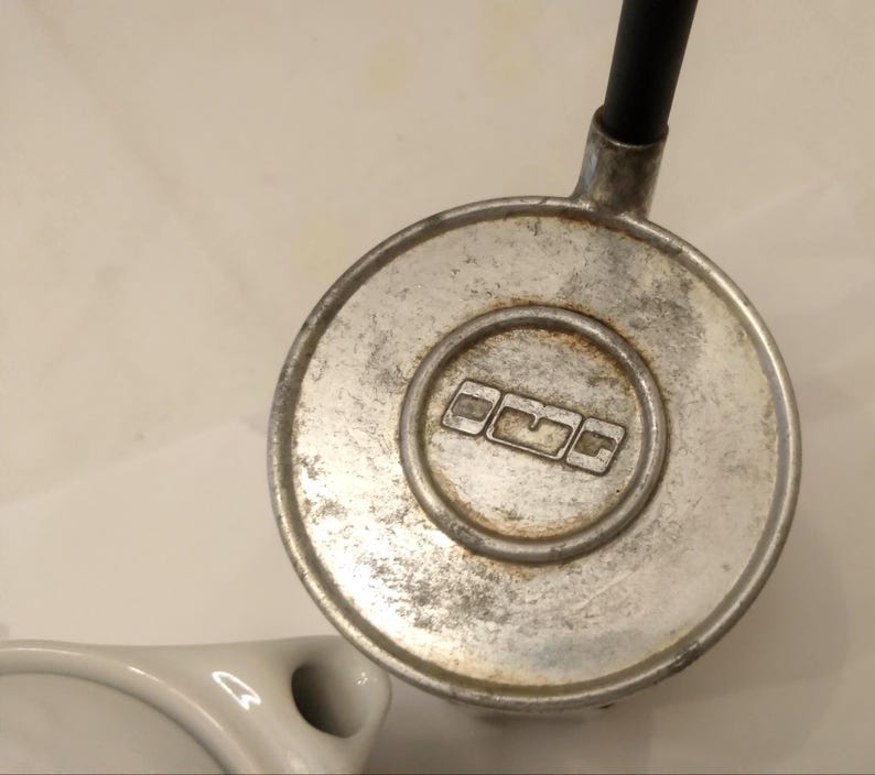 May include: Close-up of a vintage, circular metal object with a black tube attached. The object has a worn, brushed metal finish with a recessed center featuring a logo. A white ceramic object is visible in the lower left corner.