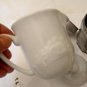 May include: A white ceramic creamer with a handle and a small spout. The creamer is slightly tilted, revealing its interior. A matching lid and a metal coffee maker are also visible.