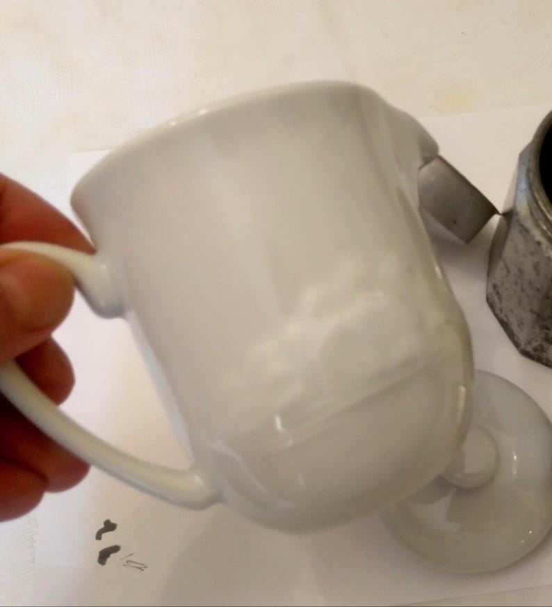 May include: A white ceramic creamer with a handle and a small spout. The creamer is slightly tilted, revealing its interior. A matching lid and a metal coffee maker are also visible.