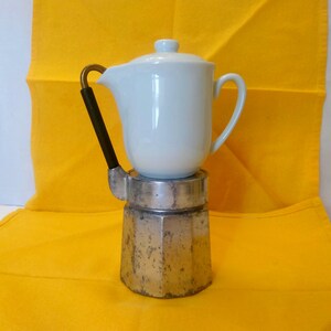 May include: A white ceramic coffee pot with a lid sits atop a silver and metal coffee maker. The coffee pot has a black handle with a gold accent. The background is a bright yellow cloth.