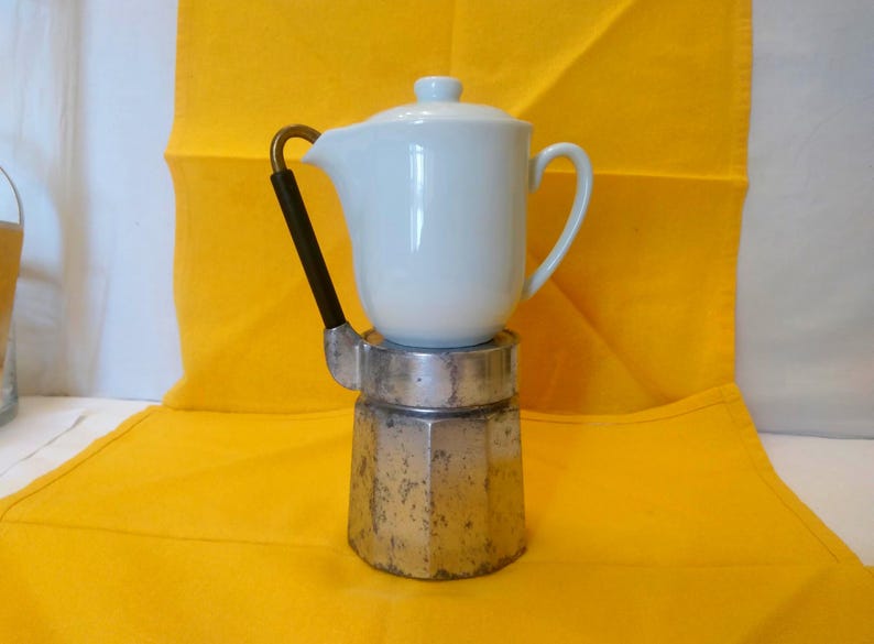 May include: A white ceramic coffee pot with a lid sits atop a silver and metal coffee maker. The coffee pot has a black handle with a gold accent. The background is a bright yellow cloth.