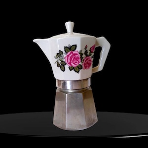 Puede incluir: Una cafetera italiana con una parte superior de cerámica blanca decorada con rosas rosadas y hojas verdes. La base está fabricada en aluminio de color plateado con una forma octogonal facetada y una tapa a juego.