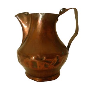 May include: A copper jug with a rounded body, a wide spout, and a curved handle attached to the side. The surface of the metal shows a hammered texture with variations in colour, ranging from deep reddish-brown to darker, oxidised tones.