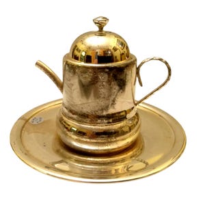 Puede incluir: Una pequeña tetera dorada con tapa y asa a juego, colocada sobre un platillo redondo. La tetera tiene un pico y un pomo decorativo en la parte superior. El juego es de metal y tiene un acabado brillante.