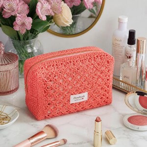 May include: A rectangular coral-coloured crochet makeup bag featuring an open-work diamond pattern and a gold-tone zip. A small white rectangular label is sewn onto the front with the text "Handmade WITH LOVE" in black ink.