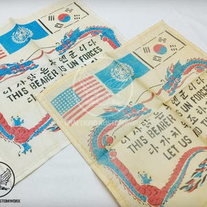 May include: Two off-white fabric squares with a vintage design. Each features a red, blue, and white dragon design, the American flag, the South Korean flag, and text in English and Korean. The text reads "THIS BEARER IS UN FORCES".