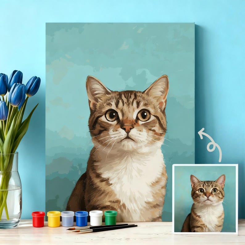 May include: A canvas print of a tabby cat with brown and white fur, set against a light blue background. The cat has large, expressive eyes. A smaller, framed image of the cat is on the right. Paint pots and brushes are in the foreground.