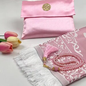 May include: A pink prayer mat with a white fringe, a matching satin pouch with a gold embellishment, and a pink beaded rosary. Artificial pink and yellow tulips are in the foreground.