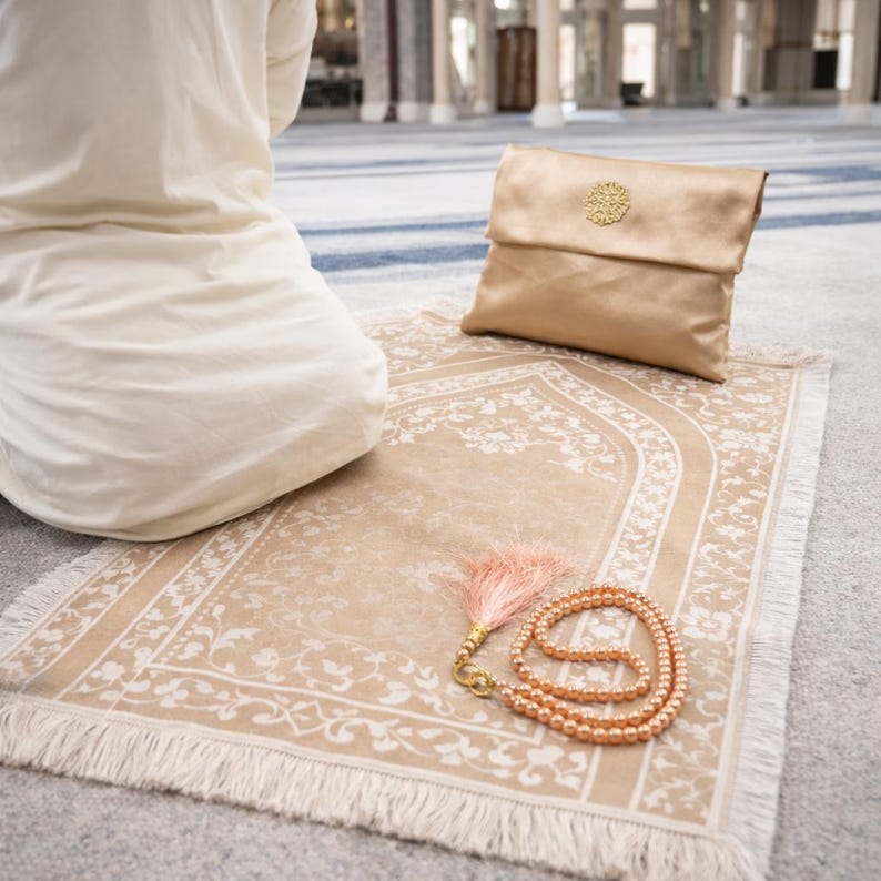May include: A beige prayer rug with a floral design and fringe, a gold satin pouch with a decorative emblem, and a string of peach-colored prayer beads with a tassel. The items are arranged on a light gray surface.