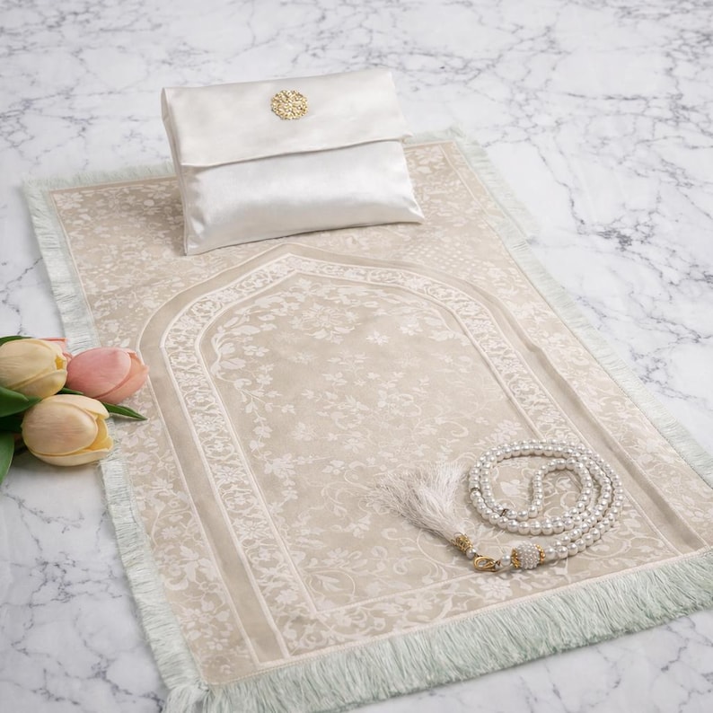 May include: A beige prayer rug with a floral pattern, a white satin pouch with a gold embellishment, and a white beaded prayer beads. The rug has a fringed edge. The items are arranged on a marble surface.