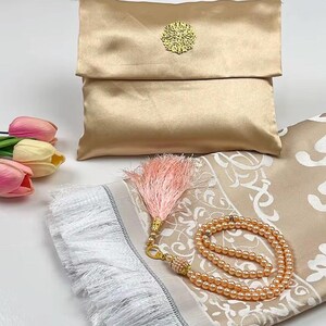 May include: A gold-colored prayer mat with a matching satin pouch and prayer beads. The mat has a beige floral design and white fringe. A pink tassel and a bouquet of tulips are also present.