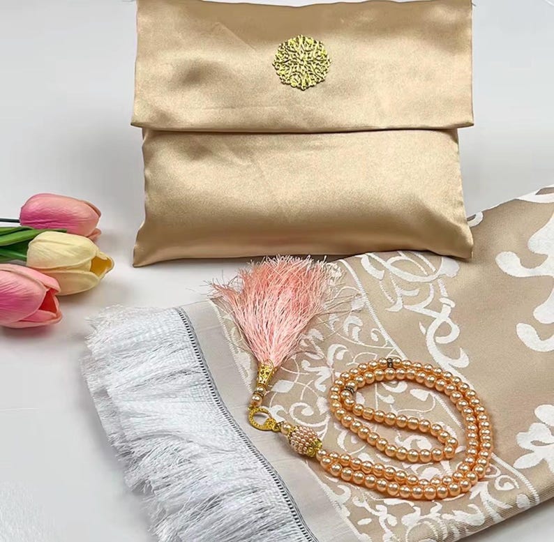 May include: A gold-colored prayer mat with a matching satin pouch and prayer beads. The mat has a beige floral design and white fringe. A pink tassel and a bouquet of tulips are also present.