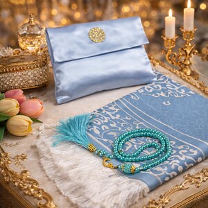 May include: A light blue satin clutch with a gold circular embellishment, a matching prayer mat with a turquoise tassel and beaded rosary. The items are arranged on a decorative surface with candles and flowers.